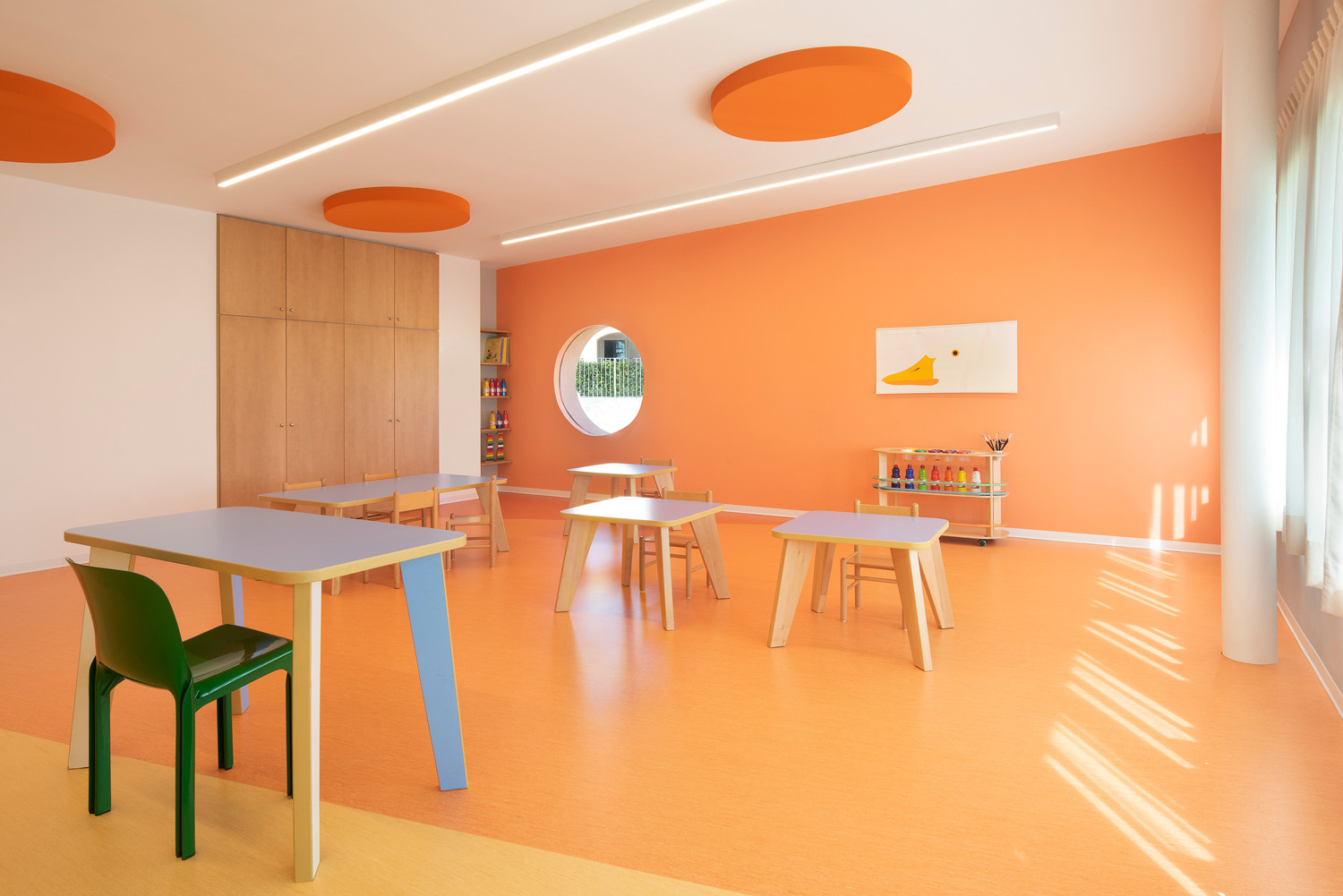 A modern, bright classroom with orange walls and floor. There are a few chairs and tables in the room. 