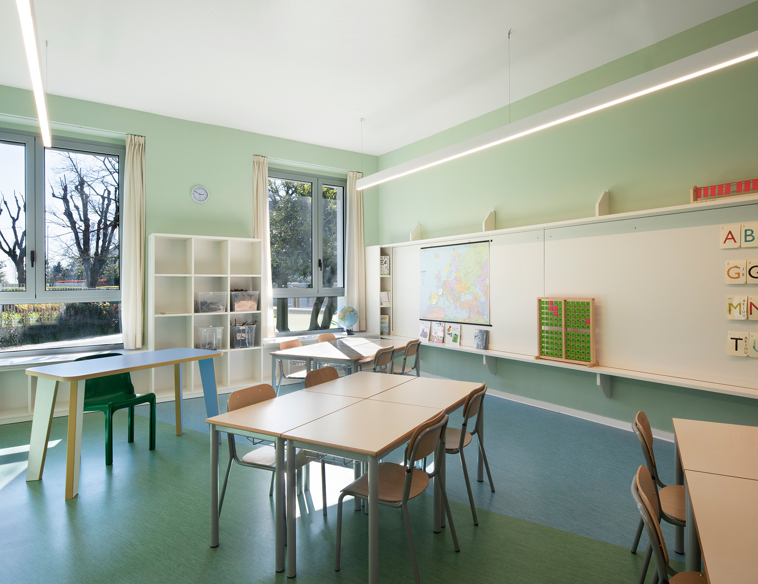 A modern classroom with a large whiteboard, green walls and tables and chairs, as well as large windows offering a view of the greenery.