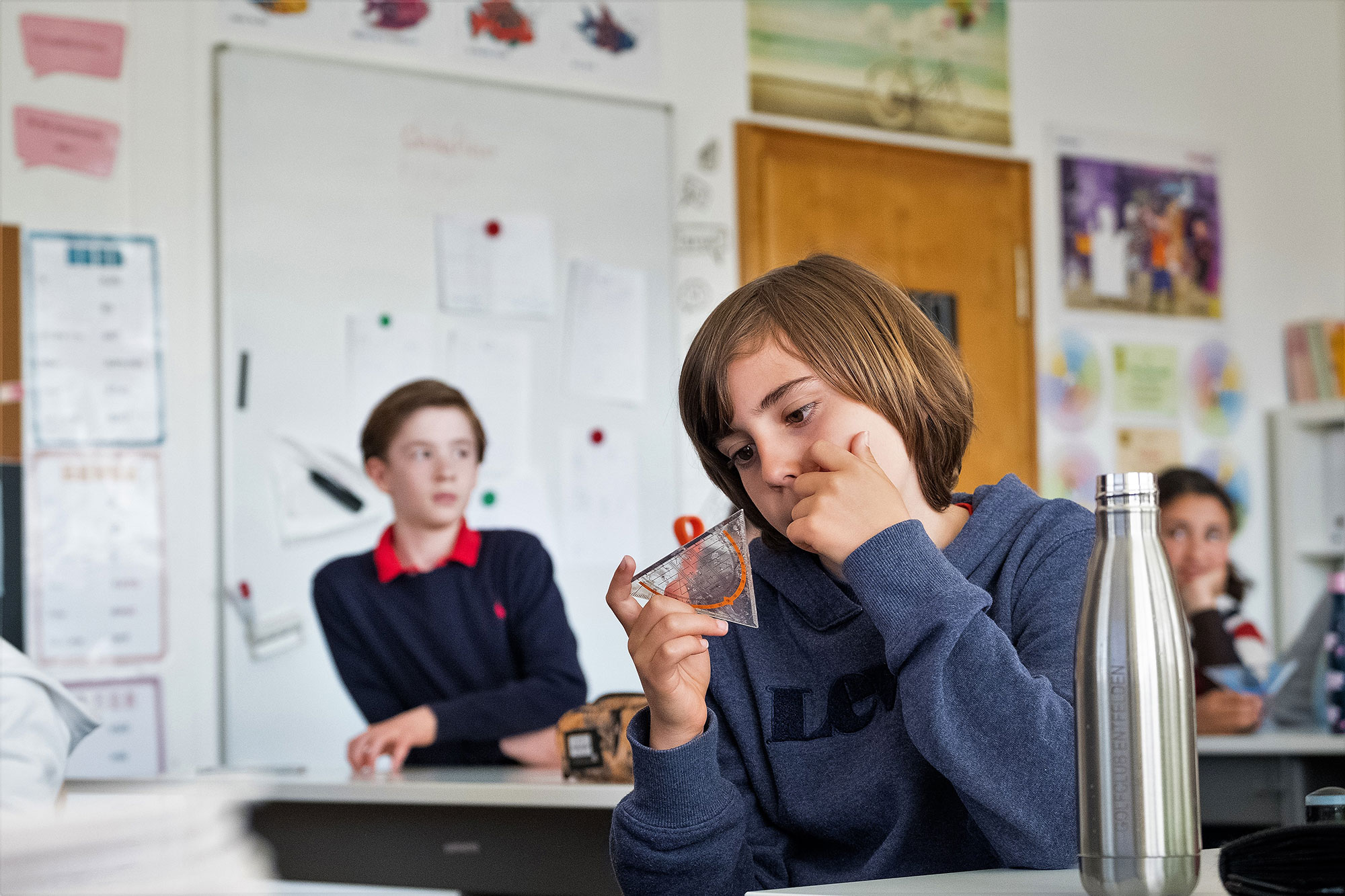 A primary school boy is looking intently at a set square. Two other children can be seen in the background.