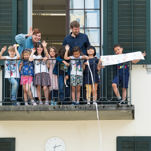 Un gruppo di bambini della scuola dell'infanzia affacciati a un balcone, insieme a due insegnanti, che salutano le persone.