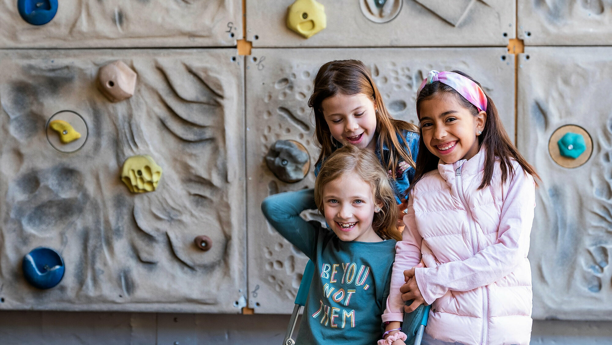 Three girls stand close together in front of the school's climbing wall.