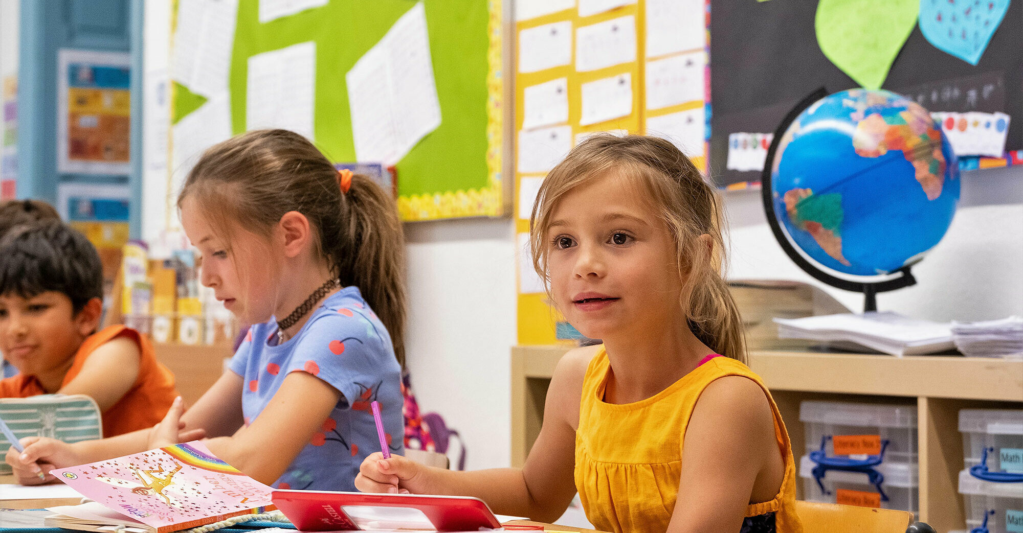 Due bambine della scuola elementare mentre scrivono sedute al banco. Una bambina guarda davanti a sé con gli occhi spalancati. Sullo sfondo si vedono un mappamondo blu e dei foglietti attaccati alla lavagna.