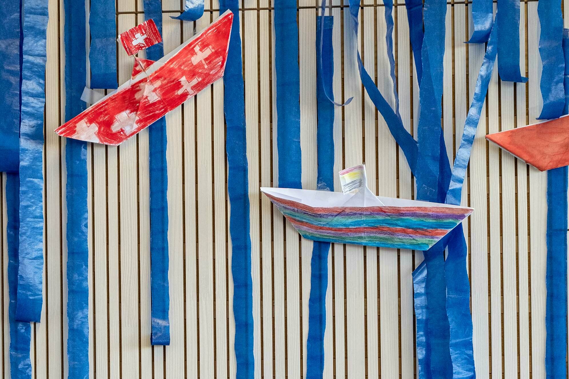 Self-made, painted paper boats hang on the wall as decoration. One of the boats is painted with the Swiss flag.