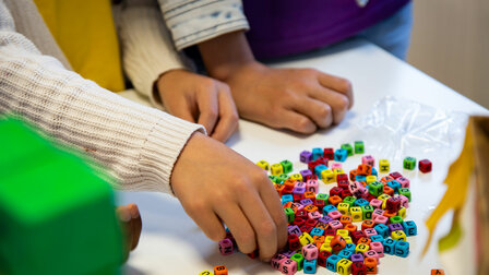Primo piano delle mani di tre bambini su un tavolo. Un bambino ha allungato il braccio per prendere delle perline colorate a forma di lettera da un mucchietto.