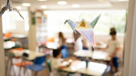 A handmade, painted origami crane hangs in the classroom, which fades into the background.