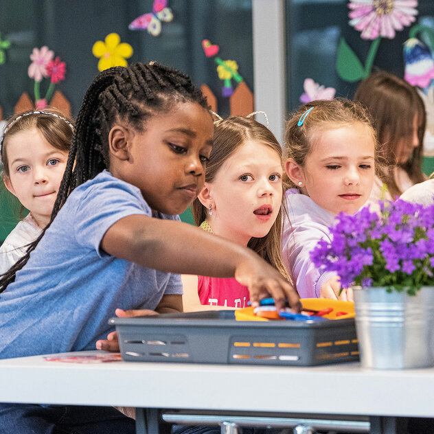 Sei bambine sedute a un tavolo. Una bambina cerca una matita da una scatola grigia. Si vedono due piccoli vasi di fiori.