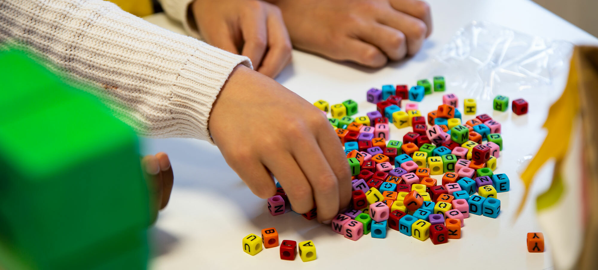 Primo piano delle mani di tre bambini su un tavolo. Un bambino ha allungato il braccio per prendere delle perline colorate a forma di lettera da un mucchietto.