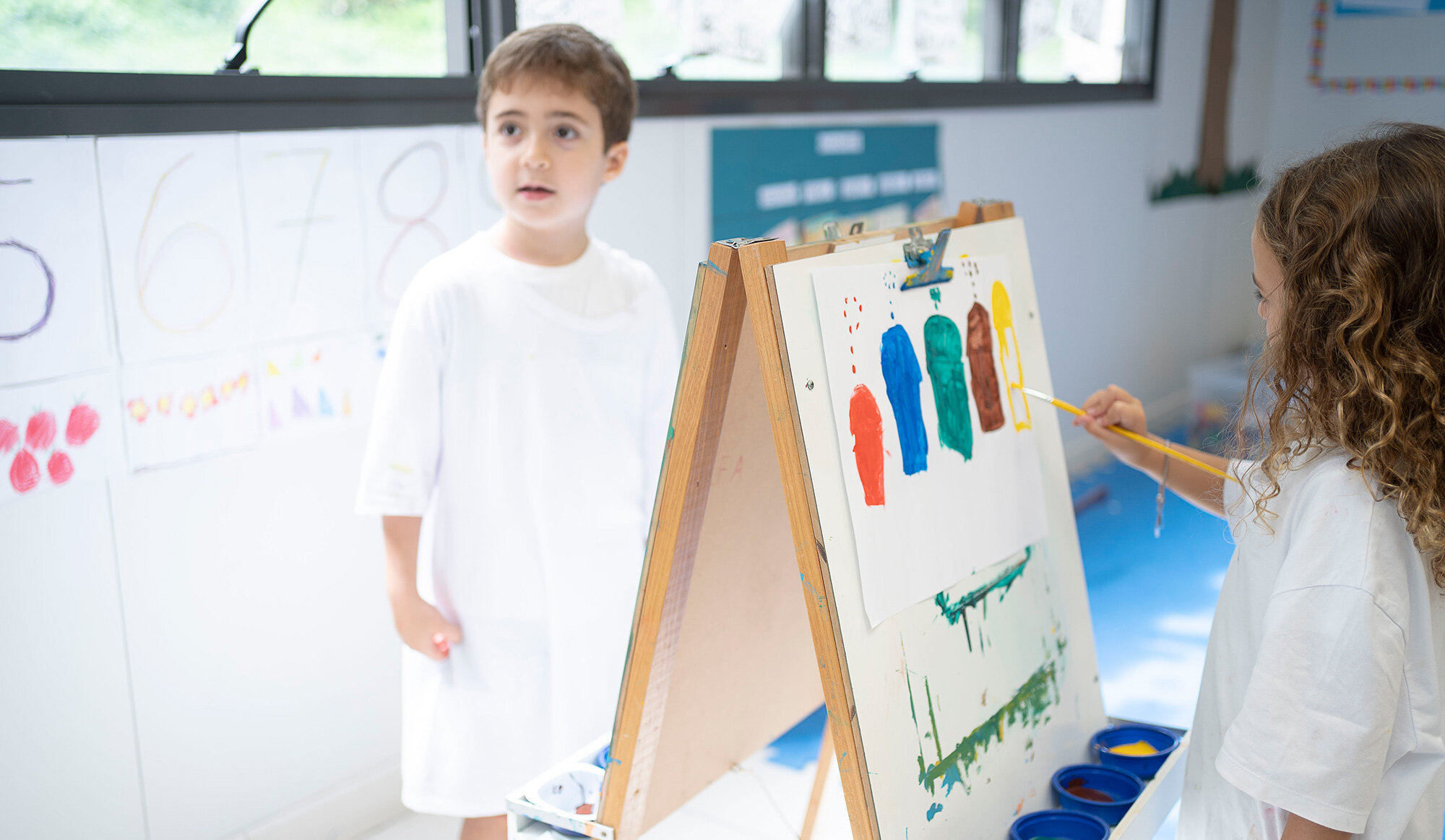 Una bambina che indossa un grembiule da pittura dipinge con un pennello elementi colorati su un foglio di carta bianco appoggiato su un cavalletto.